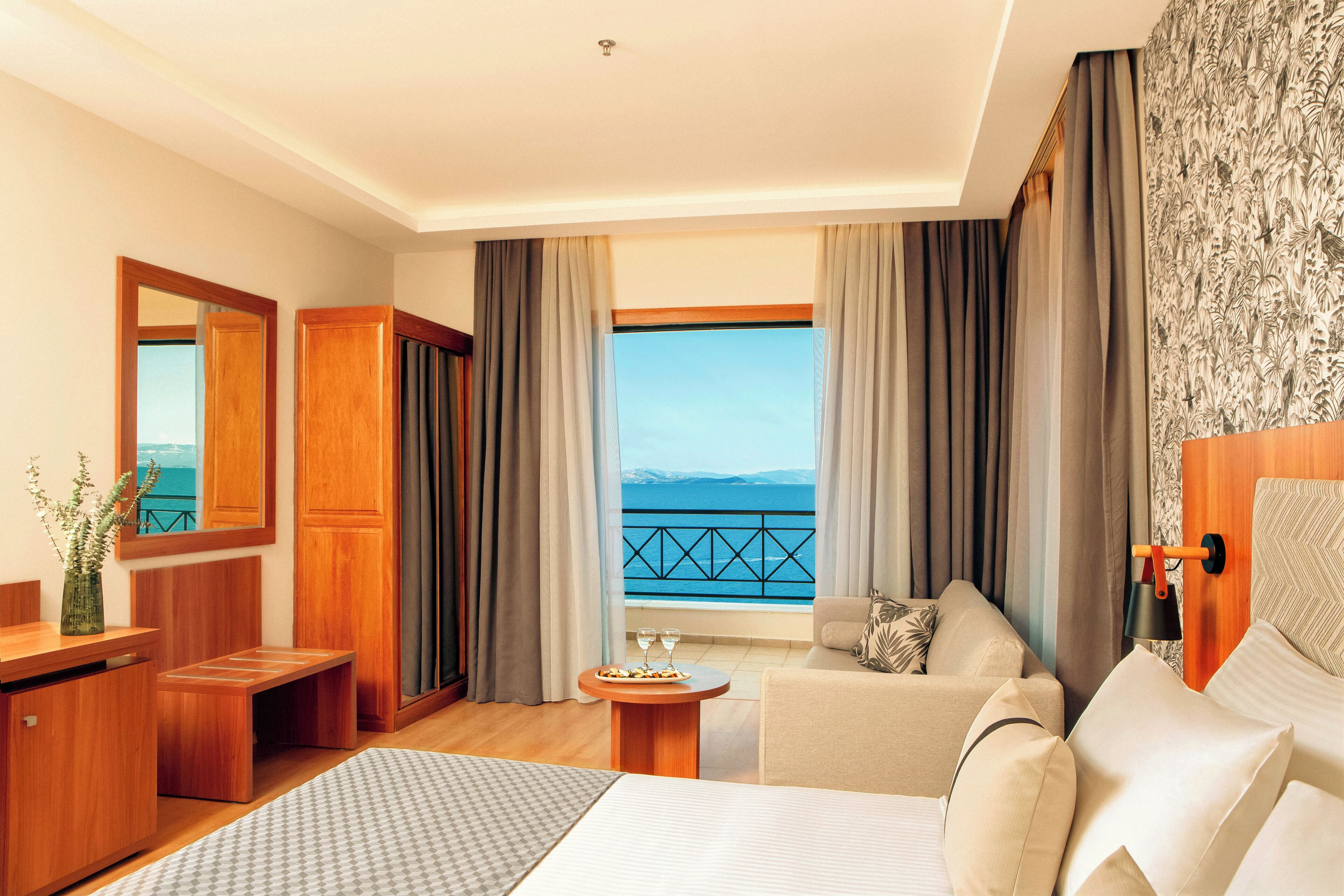 A hotel room with a bed, a sofa, a small table with a vase and two glasses, a wooden bench, and a view of the sea through large windows.