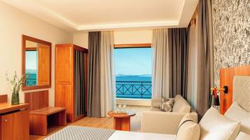 A hotel room with a bed, a sofa, a small table with a vase and two glasses, a wooden bench, and a view of the sea through large windows.