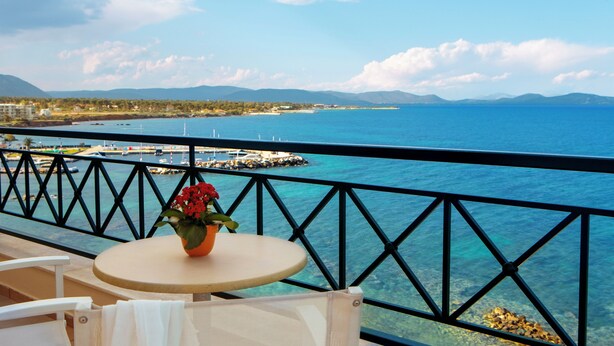 A balcony with a table and chairs, overlooking a coastal view with clear blue water, a pier, and distant mountains. - Zeus Essence Ramada by Wyndham Athens (Marathon)