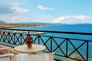 A balcony with a table and chairs, overlooking a coastal view with clear blue water, a pier, and distant mountains.