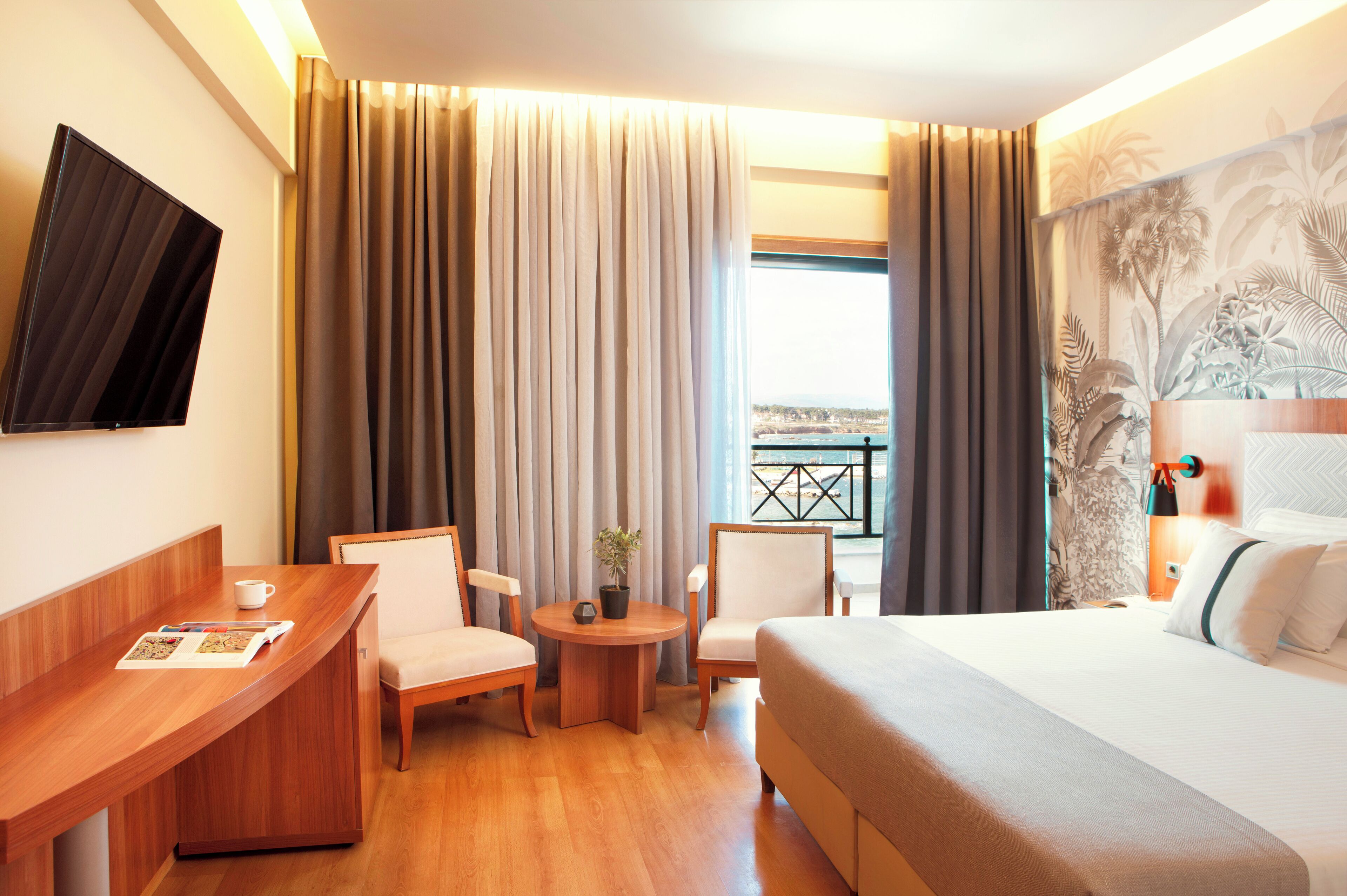A hotel room with a bed, two chairs, a small table with a plant, a coffee cup, and a magazine.
