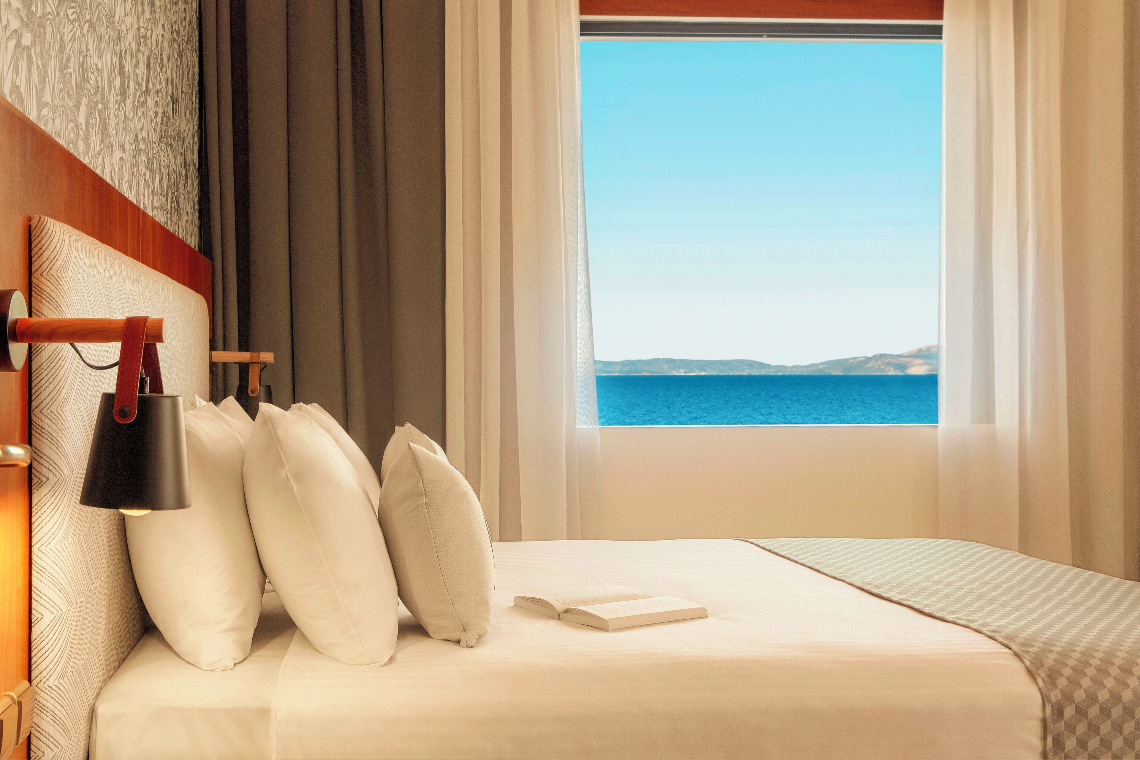 Habitación de hotel con cama, almohadas, lámpara de noche y vista al mar.