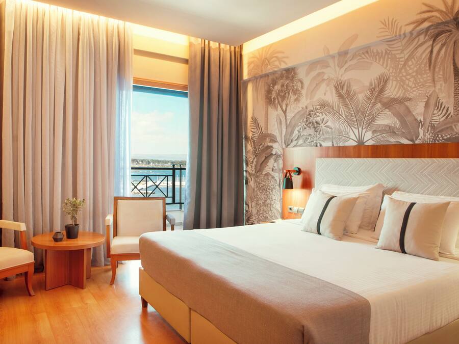 Habitación de hotel con una cama, dos sillas, una mesita con una planta y vista a un balcón con barandilla.