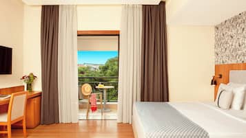 Habitación de hotel con cama, escritorio, silla, una mesita con un sombrero y un florero, y balcón con vistas a zonas verdes y edificios.
