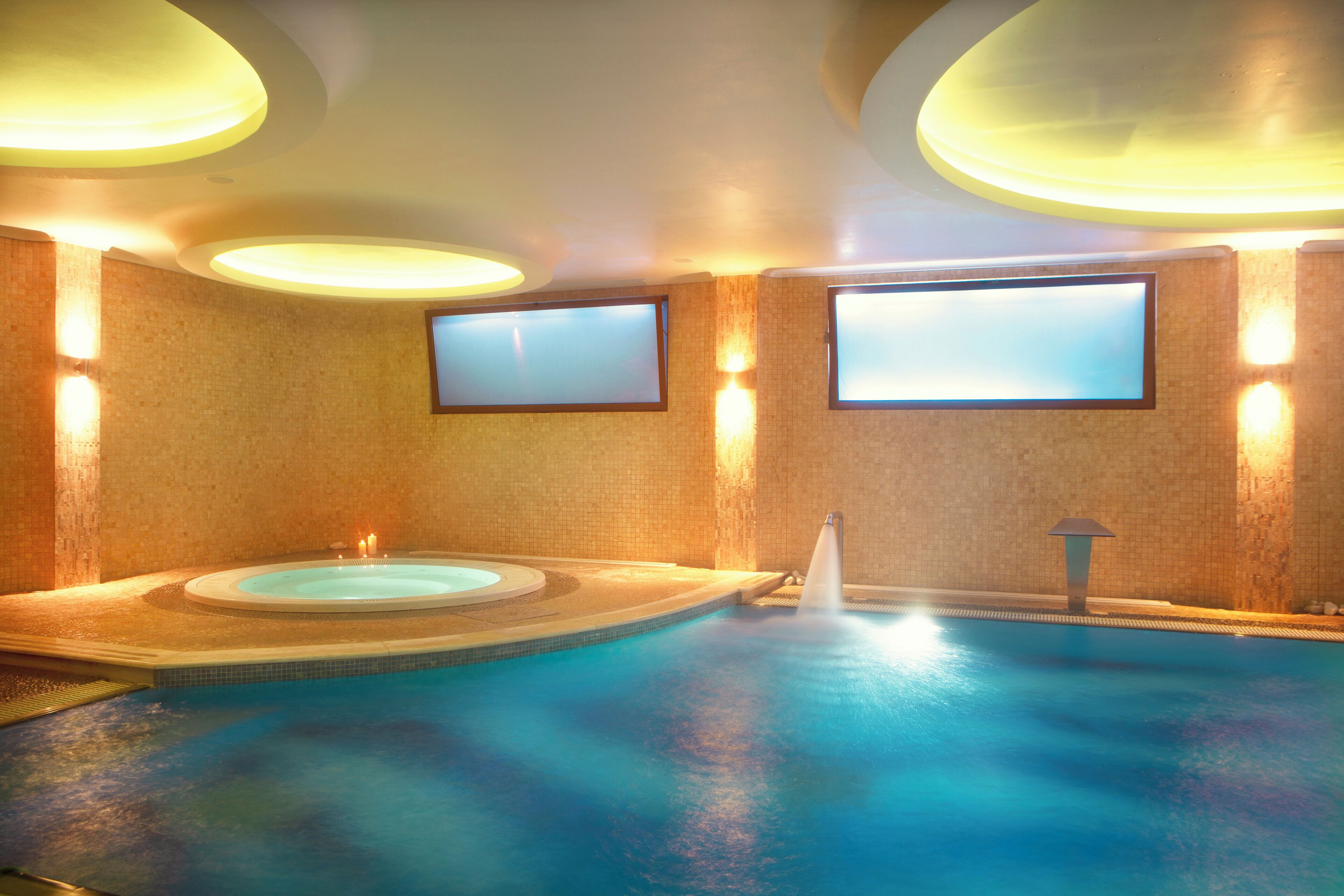 A large indoor swimming pool with a hot tub, surrounded by walls with built-in televisions.