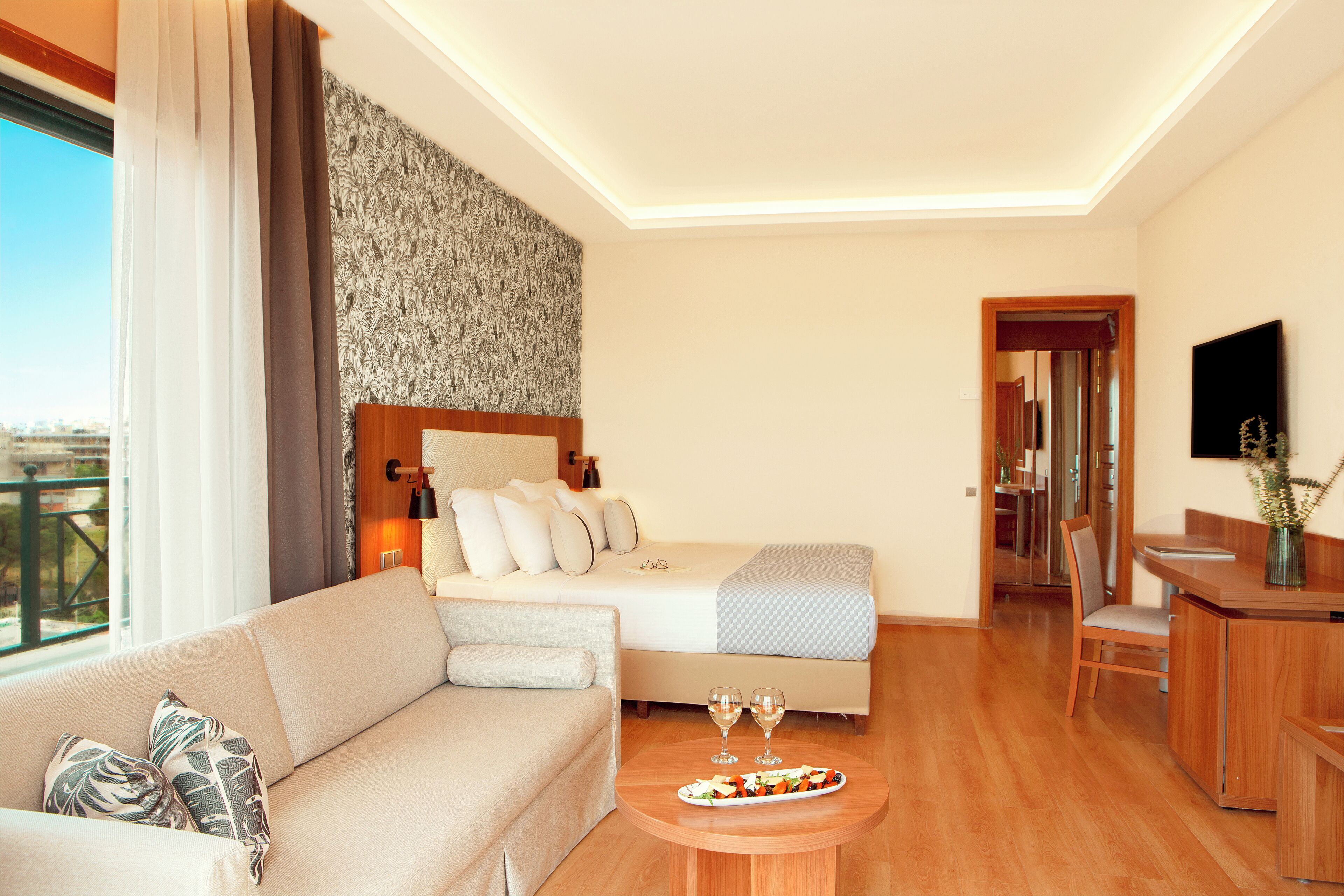 A hotel room with a bed, a sofa, a dining table with a plate of food and glasses, a TV, and a view of the outdoors.