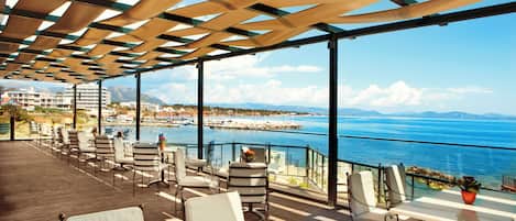 A terrace with outdoor seating, overlooking a coastal view with mountains and a marina.