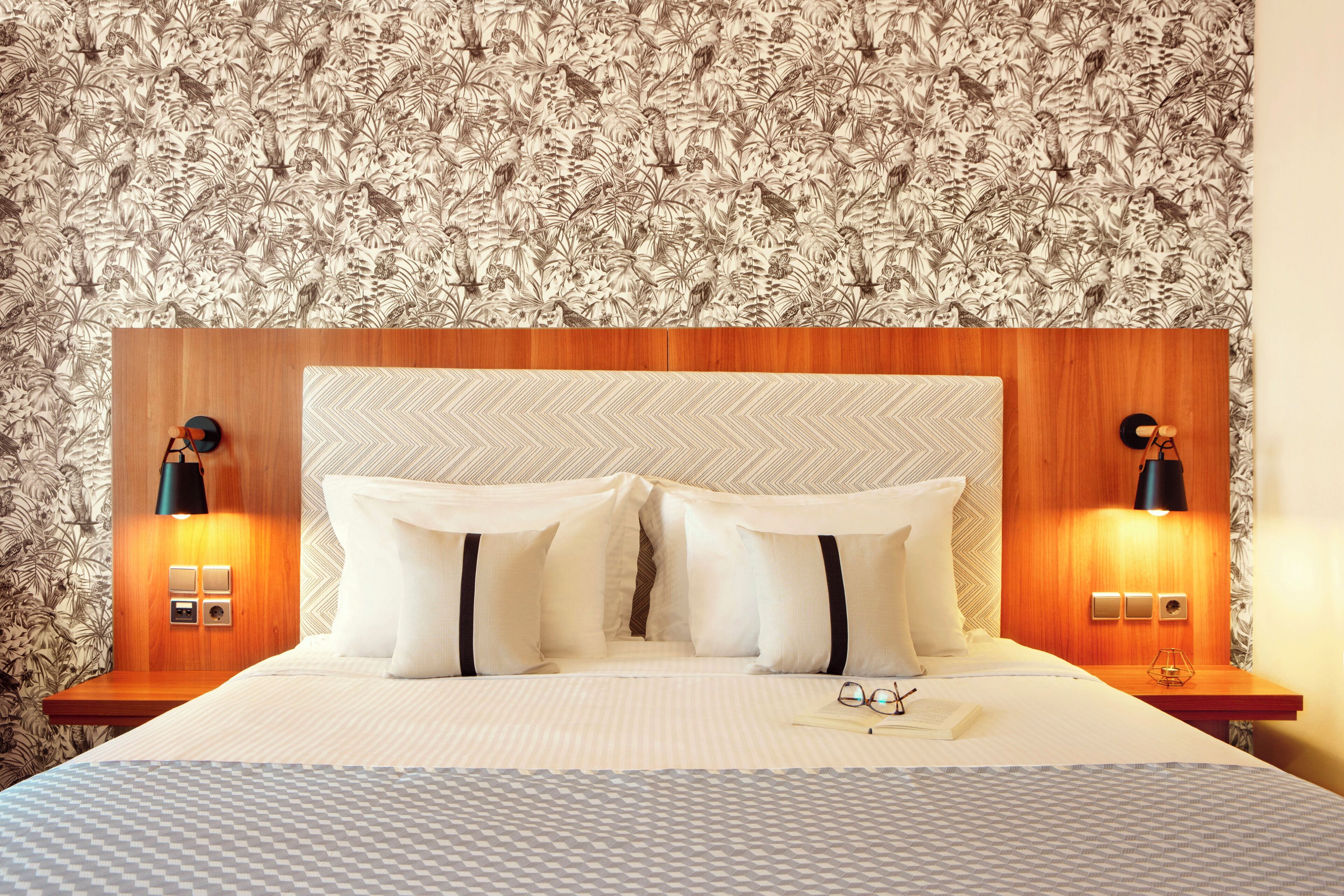 A hotel room with a patterned wallpaper, a wooden headboard, two bedside lamps, and a neatly made bed with pillows and a folded towel.