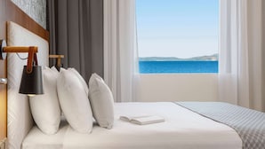 A neatly made bed with a book, white pillows, and a lamp in a room with a view of the sea. - Zeus Essence Ramada by Wyndham Athens (Marathon)