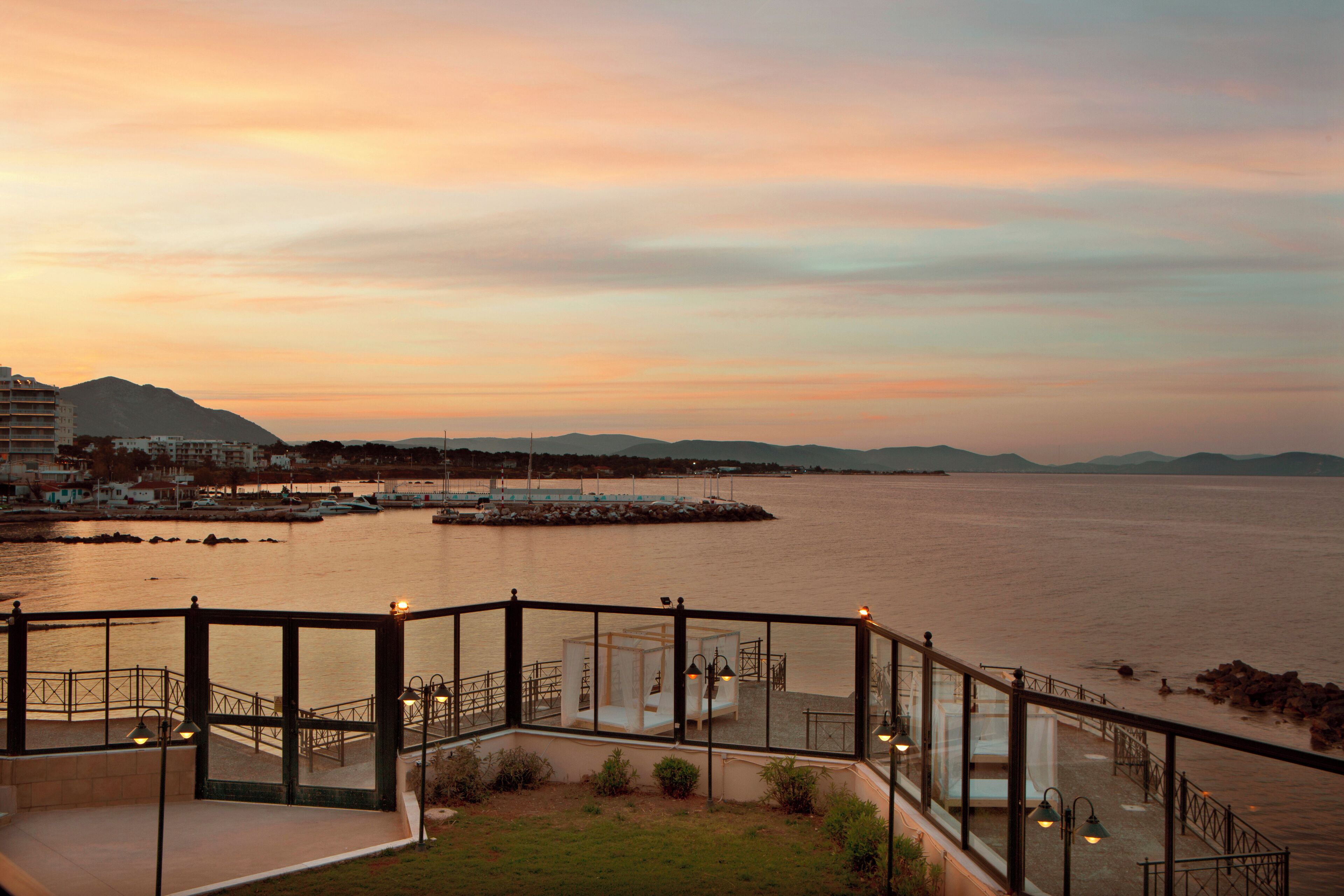 Um hotel à beira-mar com terraço envidraçado, vista para o mar e uma cadeia montanhosa ao longe.