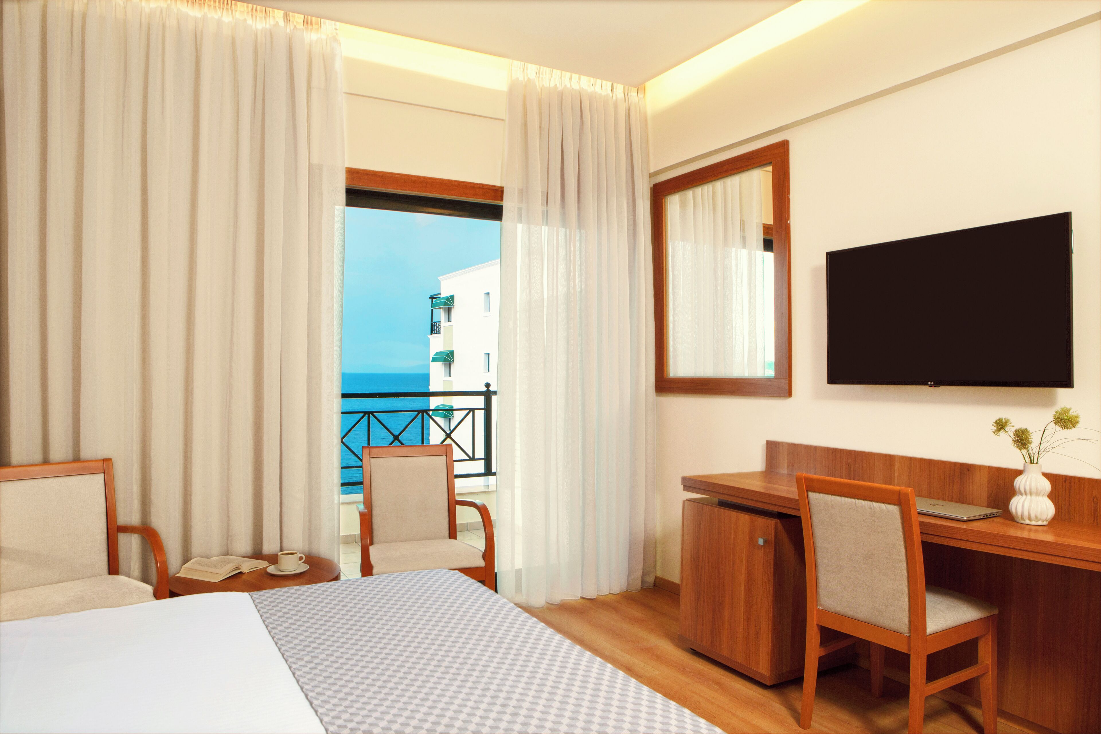 A hotel room with a bed, a desk, a chair, a TV, and a view of the sea.