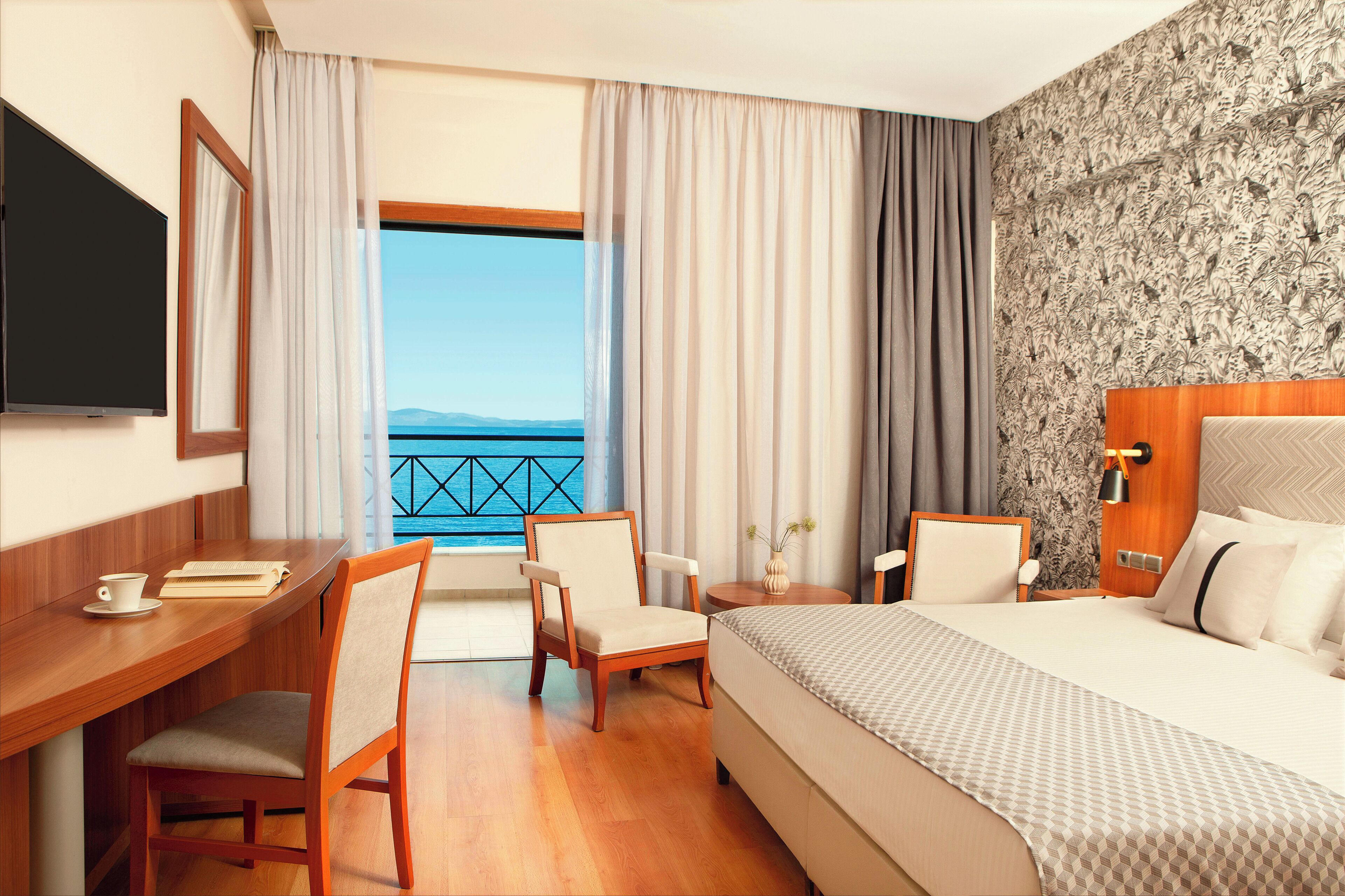 A hotel room with a bed, desk, chairs, a TV, and a view of the sea.