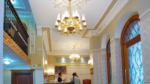 Un vestíbulo de hotel con mostrador de recepción, una araña, plantas en macetas y suelo a cuadros.