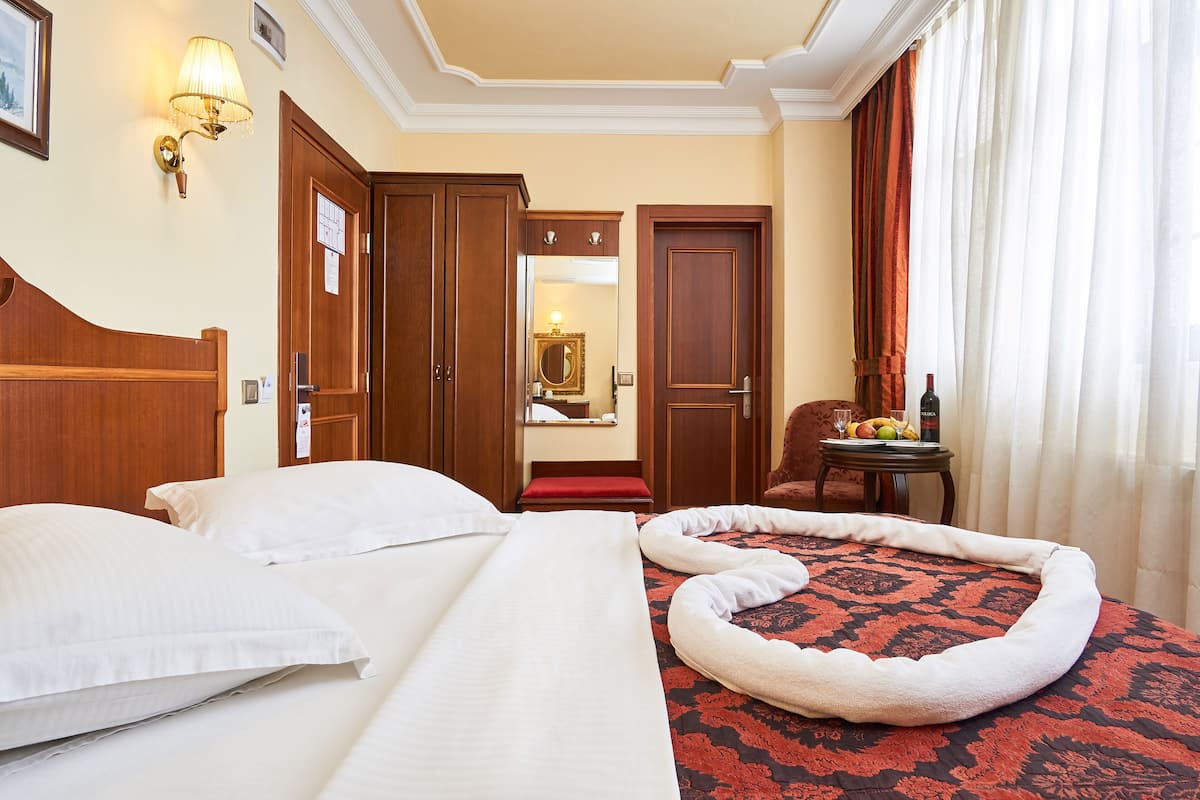 A hotel room with a bed, a red and black patterned rug, a wooden headboard, a wooden door, a mirror, and a small table with fruits and a bottle.