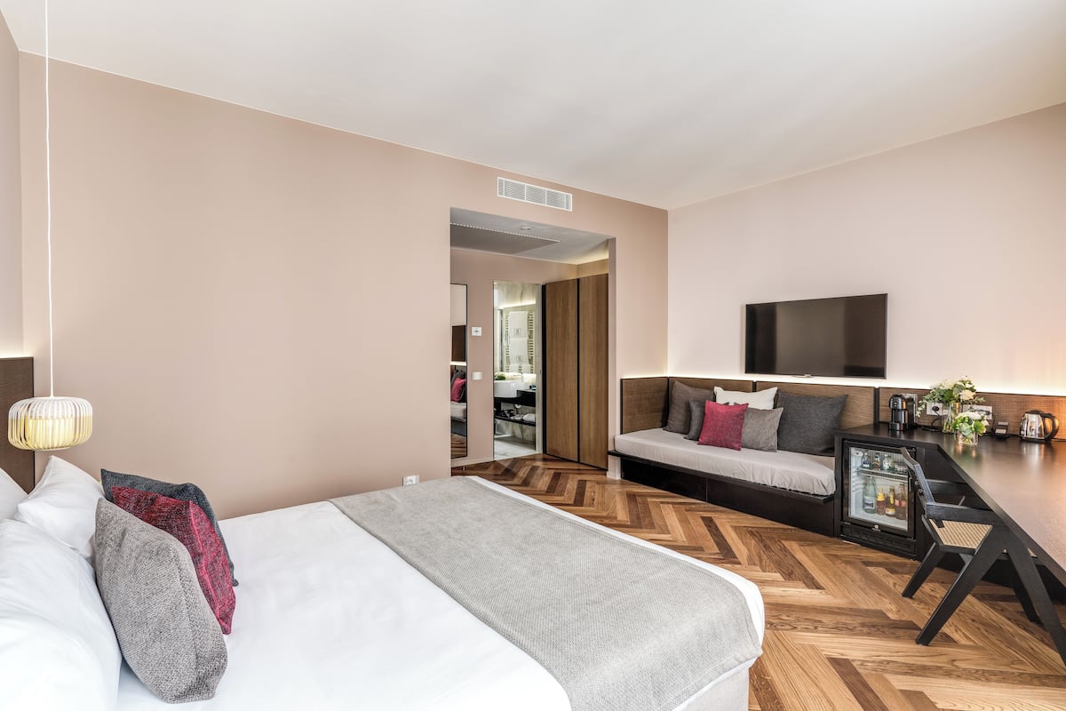 A modern hotel room with a large bed, a sofa, a dining table, and a TV mounted on the wall.