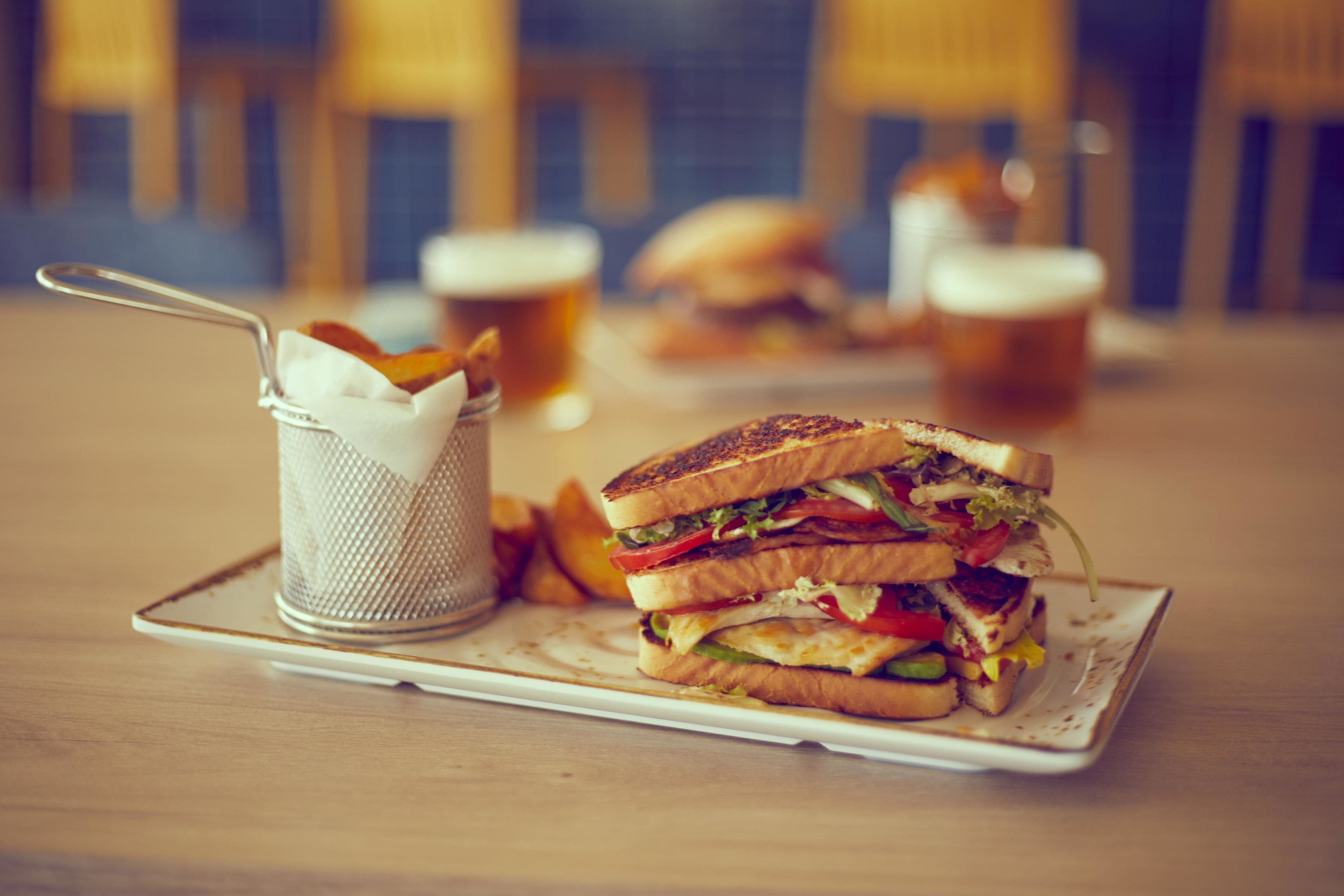 Ein Sandwich auf einem Teller mit einem Serviettenhalter und Pommes, vor unscharfem Hintergrund einer Bar.