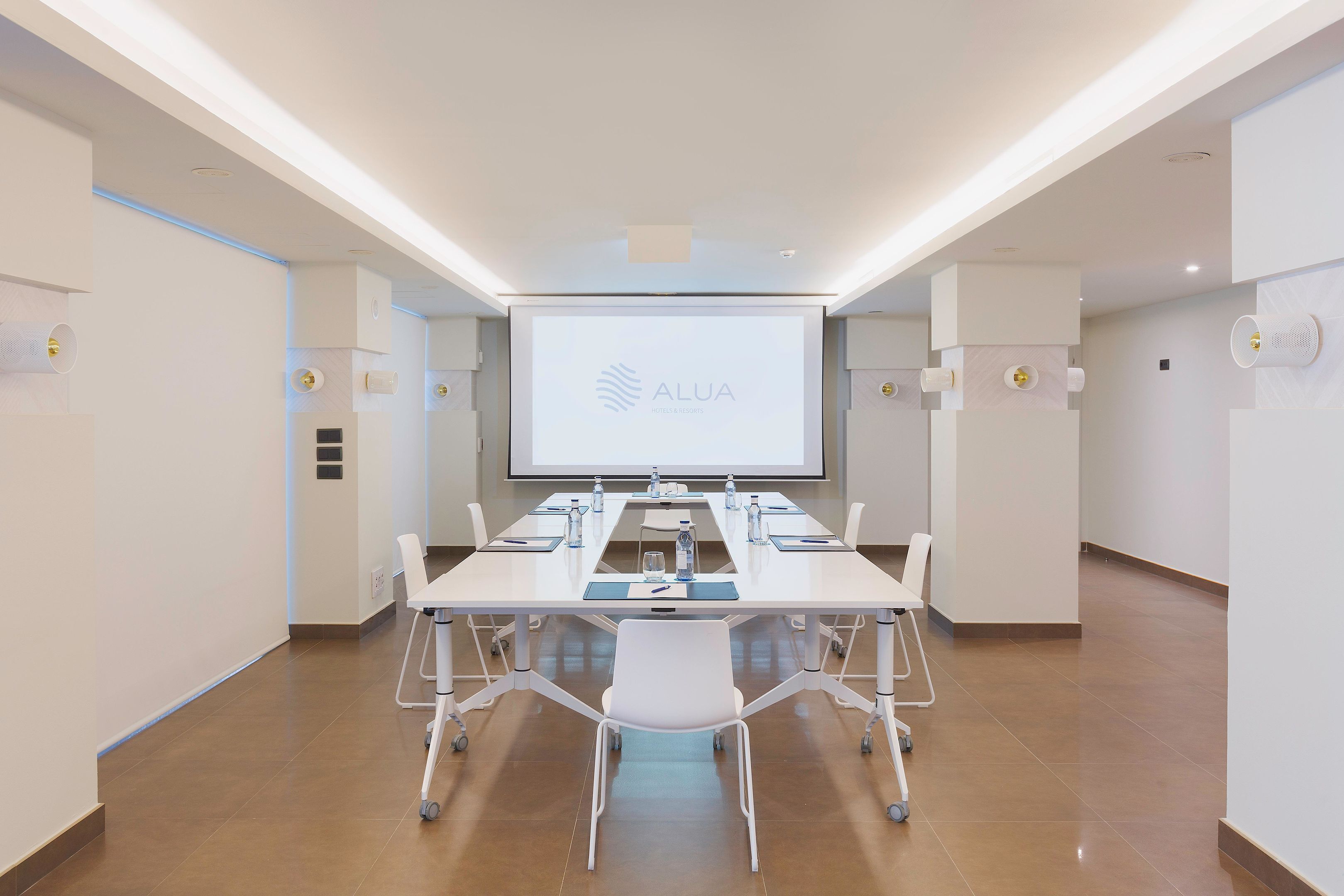 Una sala de conferències amb una taula llarga, cadires, una pantalla de projector i una presentació a la paret.