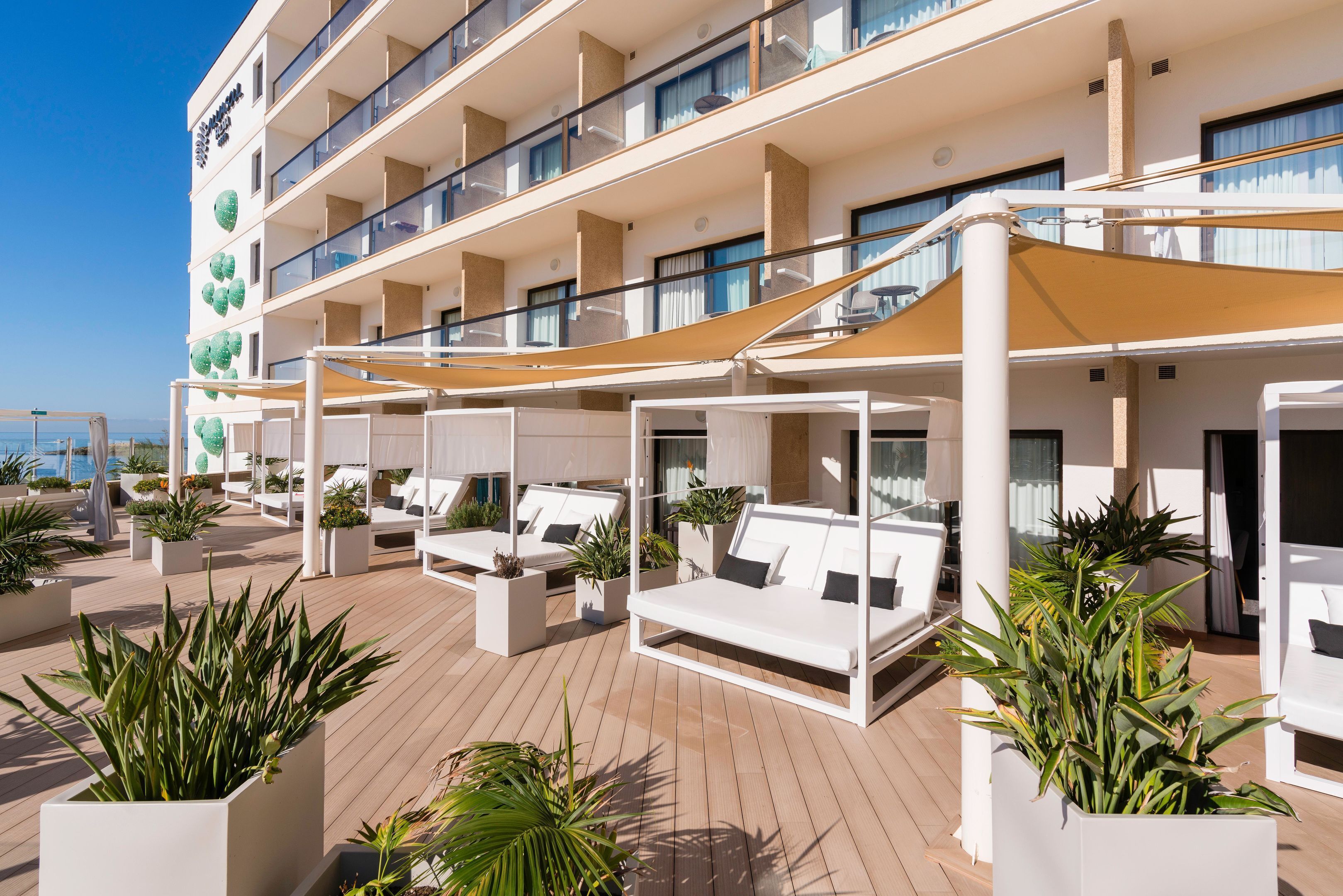 A multi-story hotel with outdoor seating areas, potted plants, and a clear view of the sea.