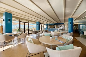 A spacious indoor area with round tables and chairs, a bar area, and large windows offering views. - AluaSoul Palma - Adults Only (Palma de Mallorca)