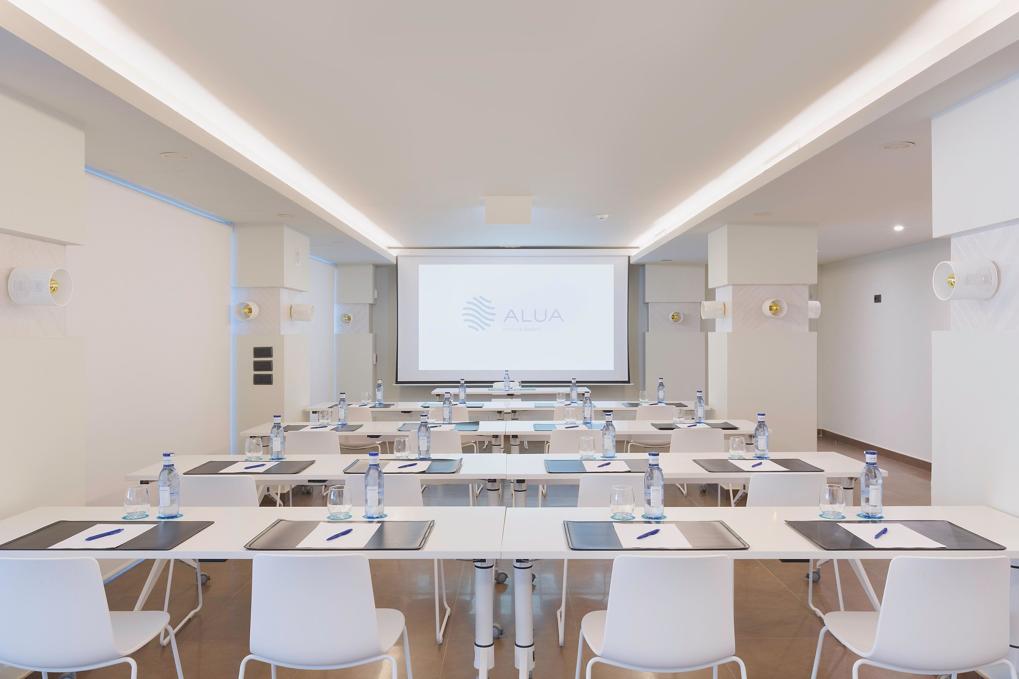 A conference room with a large screen displaying the logo 'ALUA', white tables with water bottles, and white chairs.
