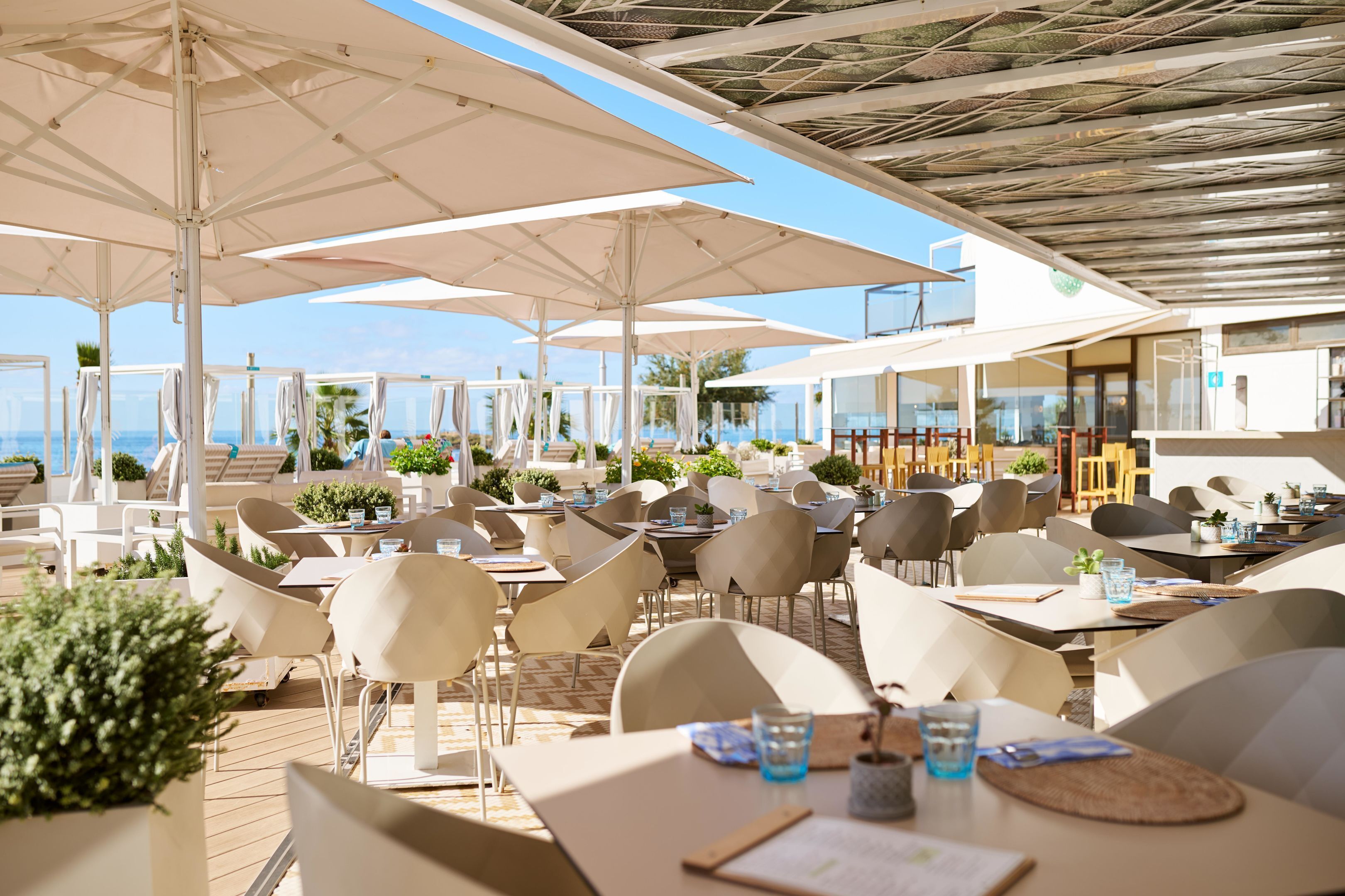A sunlit outdoor dining area with tables and chairs, umbrellas, and a view of the sea.