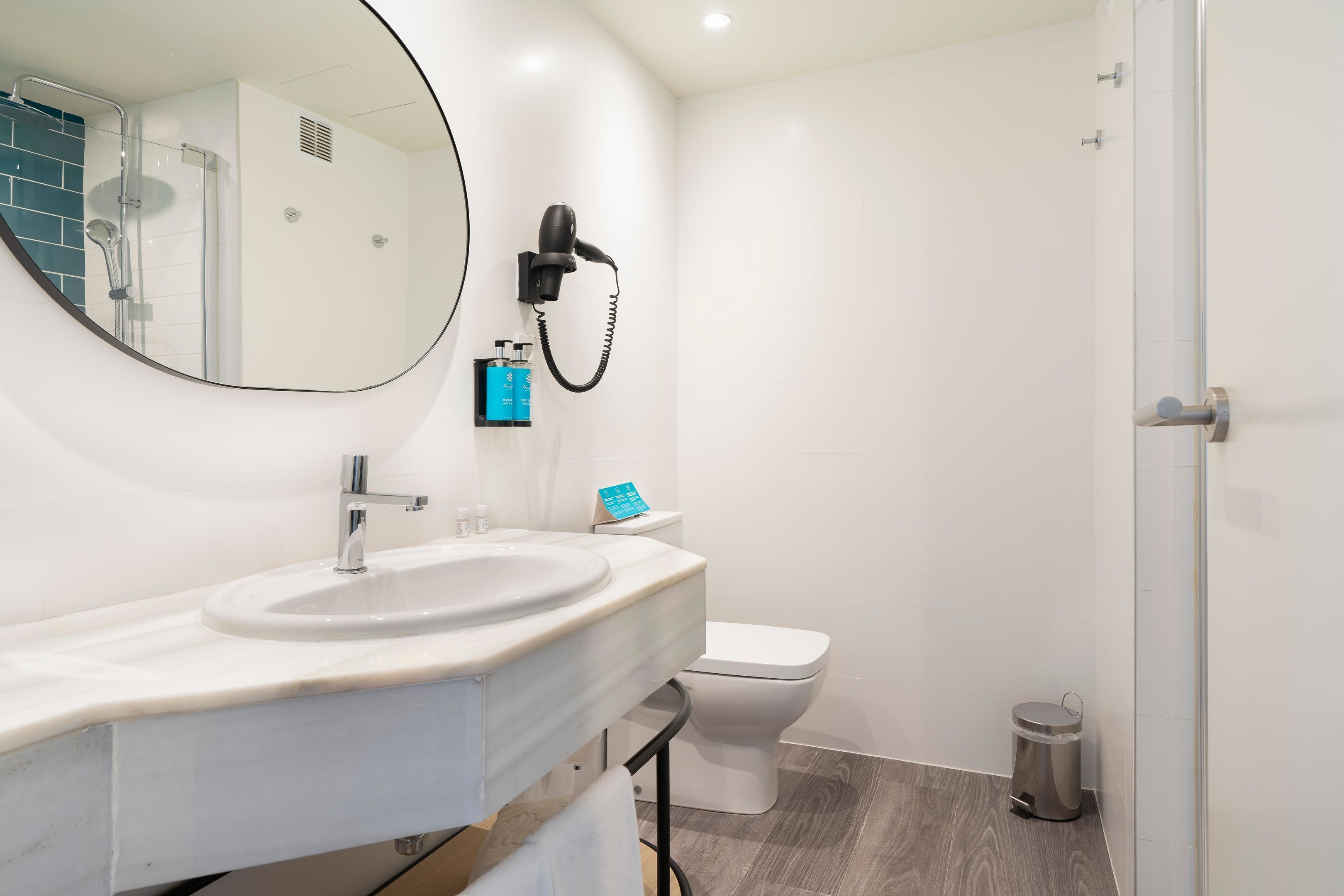 A modern bathroom with a round mirror, a sink, a toilet, and a hairdryer mounted on the wall.