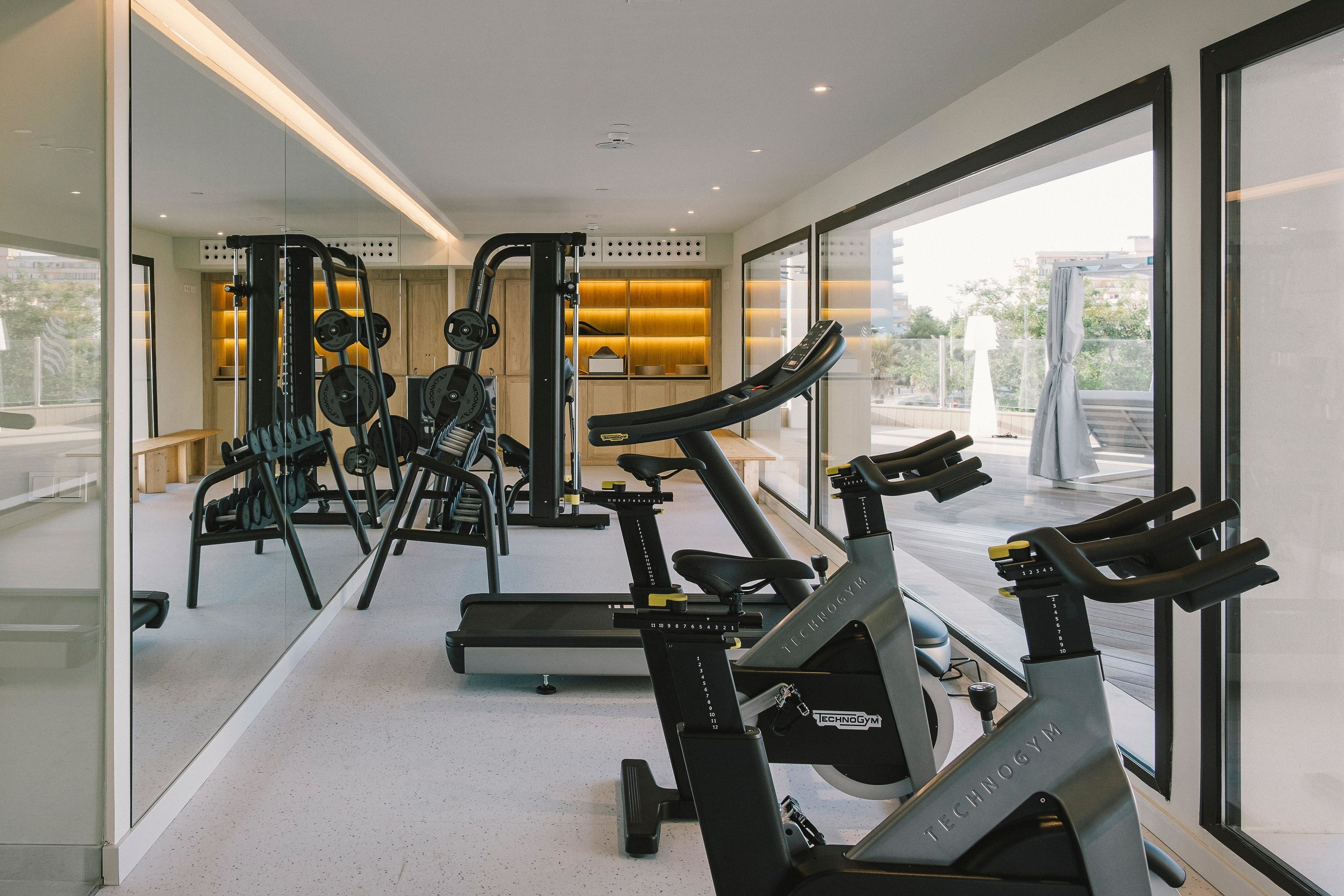 A modern gym with cardio machines, weight equipment, and a large window offering a city view.