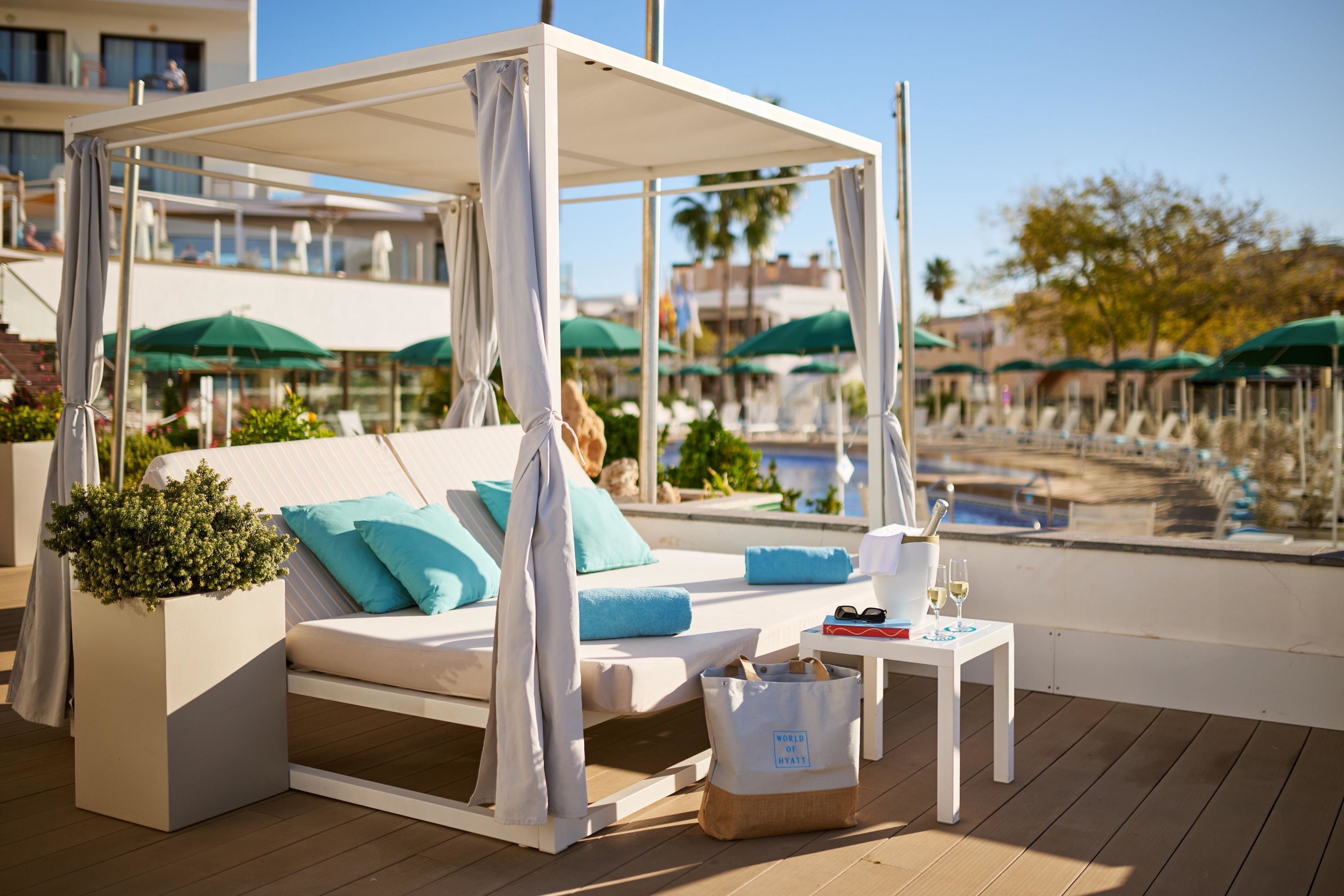 Un área de descanso junto a la piscina con una pérgola, un sofá con cojines, una mesita con una bebida y vasos, y una planta en maceta.