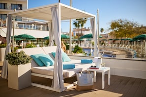 A poolside lounge area with a canopy, a sofa with cushions, a small table with a drink and glasses, and a potted plant.