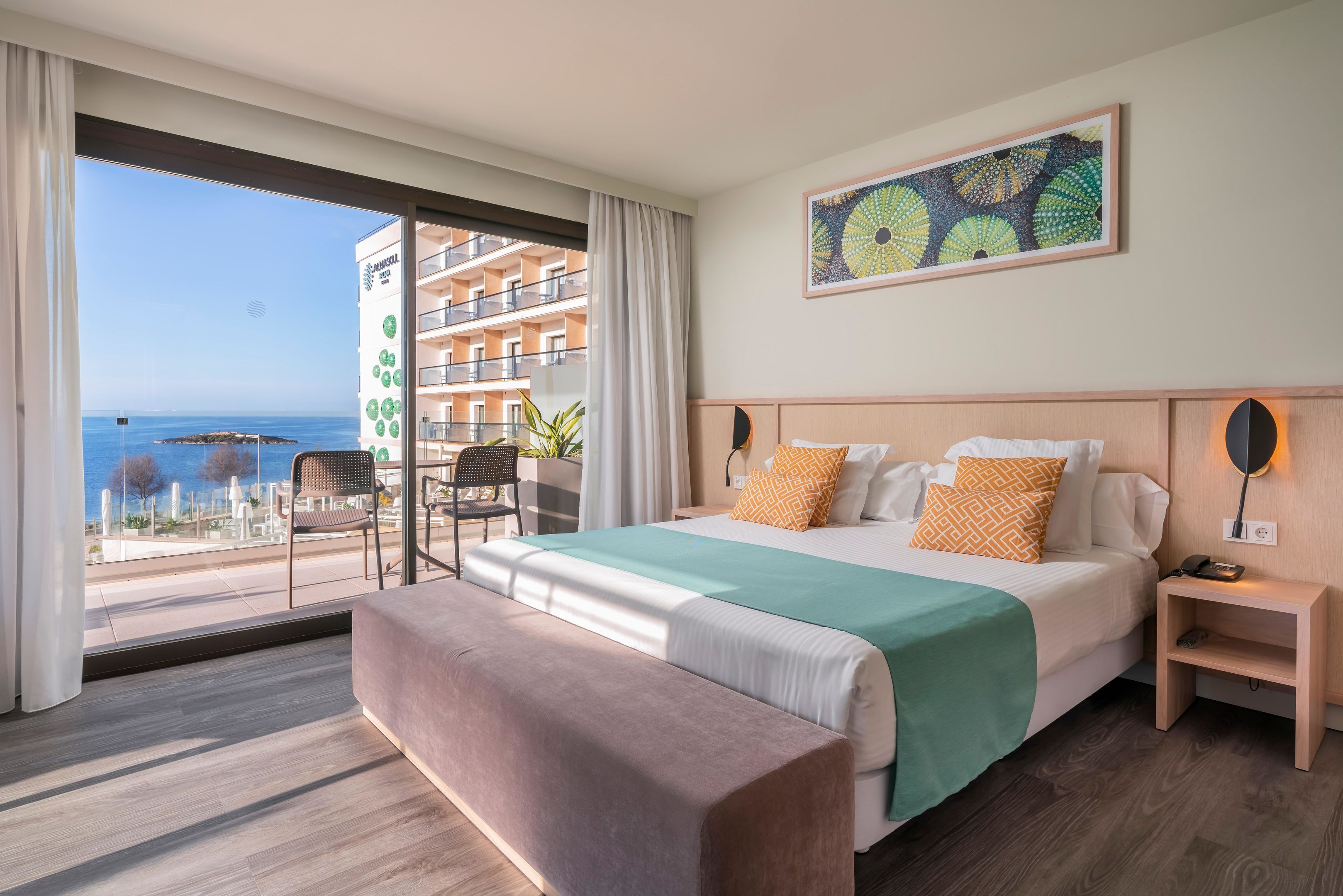 A hotel room with a large bed, a bench, a nightstand, a wall-mounted lamp, and a view of the sea through a balcony.