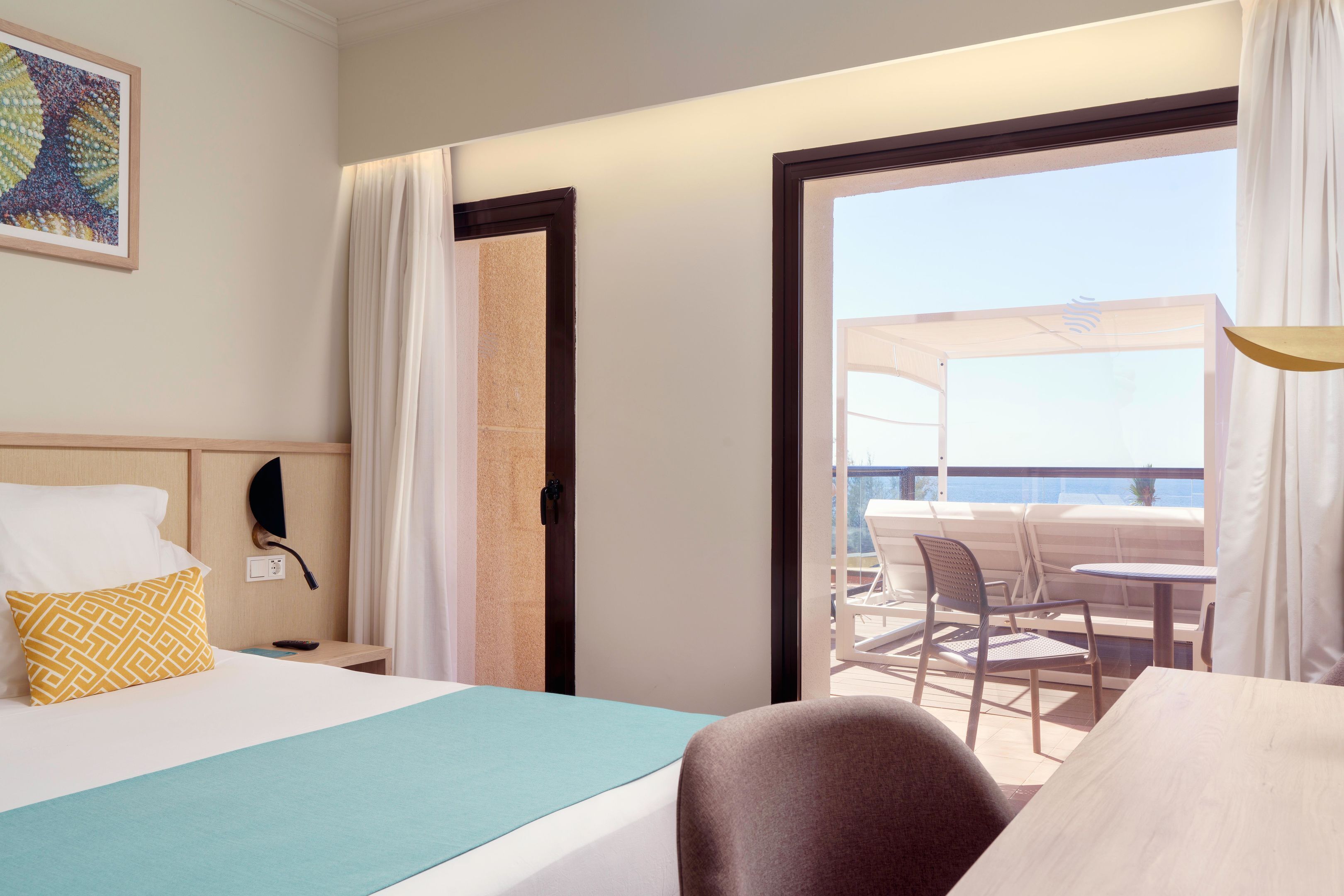 Ein Hotelzimmer mit Bett, Nachttisch, Schreibtisch, Stuhl und einem Balkon mit Sitzgelegenheit und Meerblick.