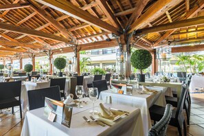 An outdoor dining area with wooden beams, tables set for two, and potted plants.