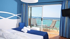 Une chambre d’hôtel avec un lit, une télévision, un balcon donnant sur la mer et une table avec des chaises.