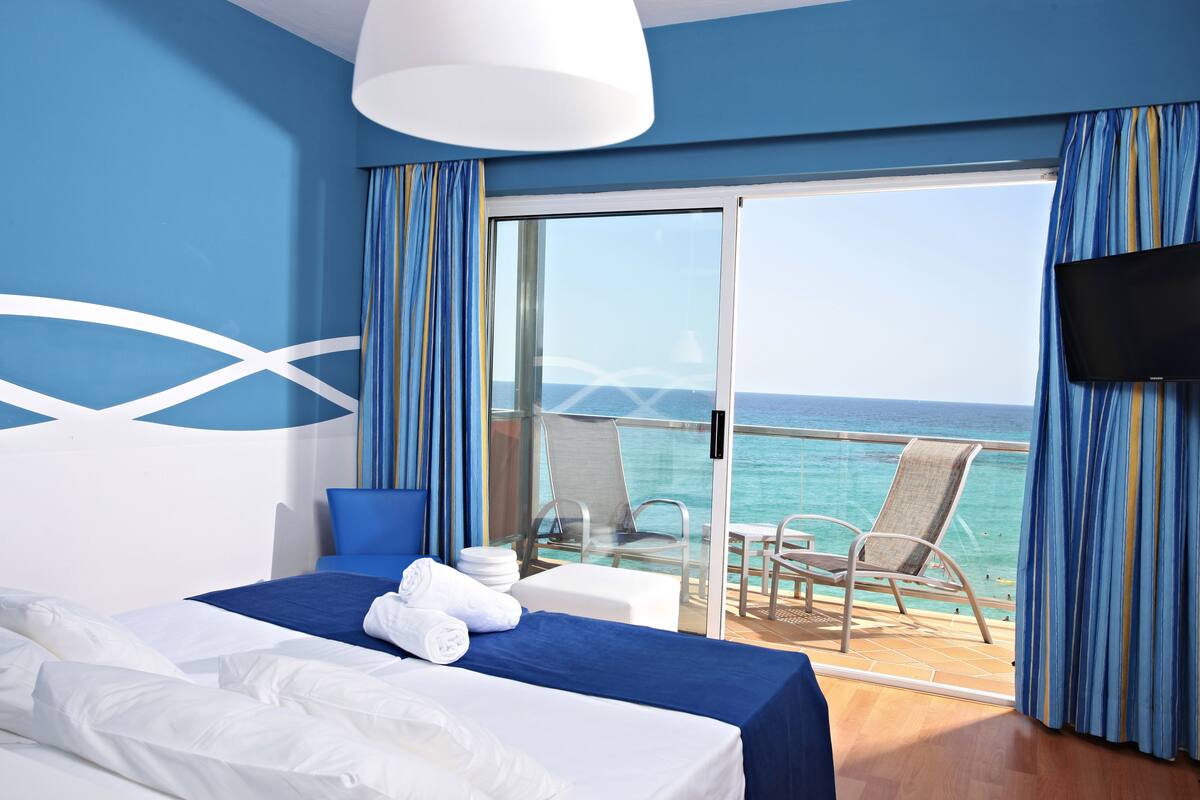 A hotel room with a bed, a TV, a balcony with a view of the sea, and a table with chairs.