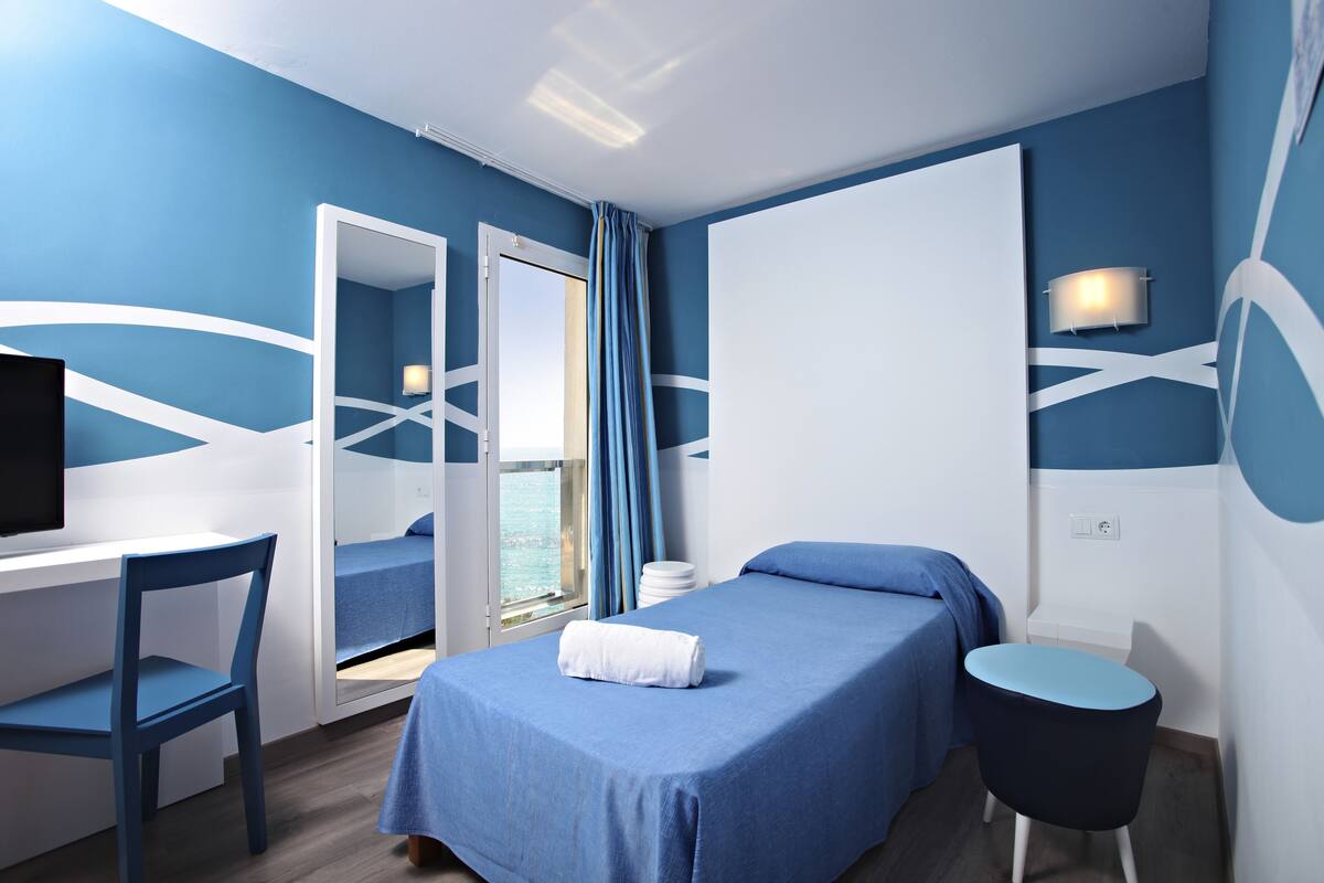 A modern hotel room with a blue and white color scheme, a bed, a desk with a chair, a small round table, and a mirror.