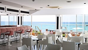 Un espace bar moderne avec des chaises blanches et roses, de grandes fenêtres offrant une vue sur l’océan et un aménagement intérieur épuré et contemporain.