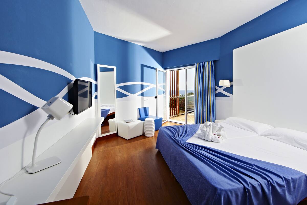 A modern hotel room with a bed, a desk with a computer, a chair, and a view of the outdoors.