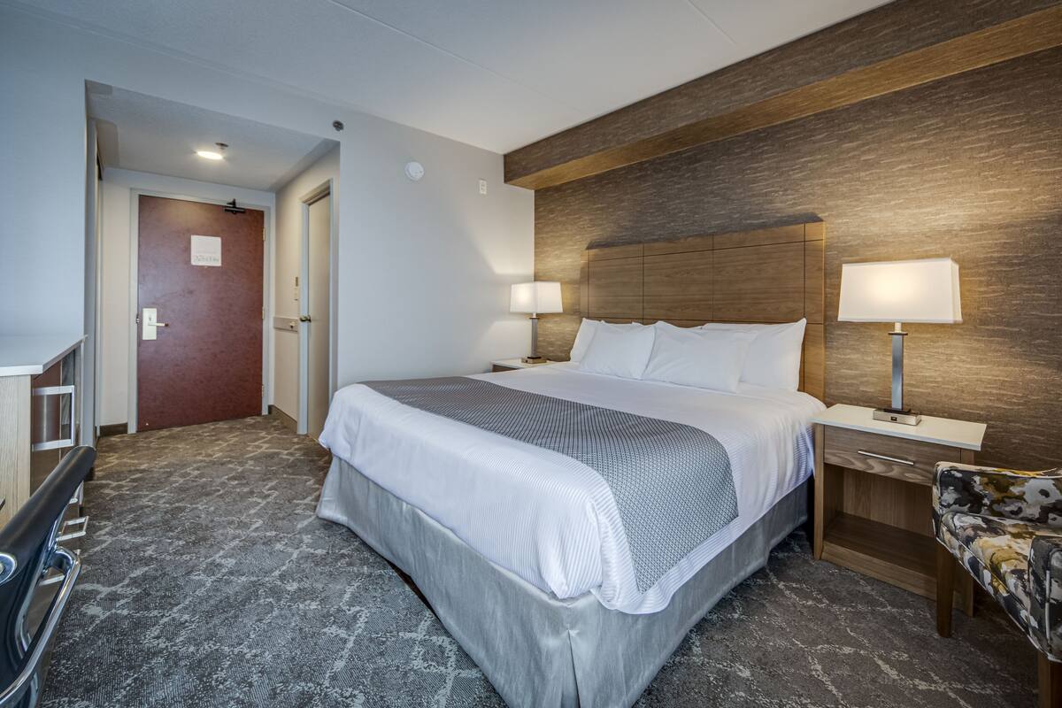 A hotel room with a large bed, two bedside tables with lamps, a floral armchair, and a wooden headboard.