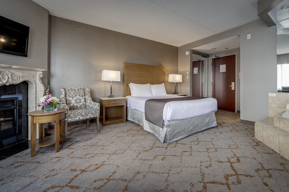 A hotel room with a large bed, two bedside tables with lamps, a chair, a fireplace, and a mirror.
