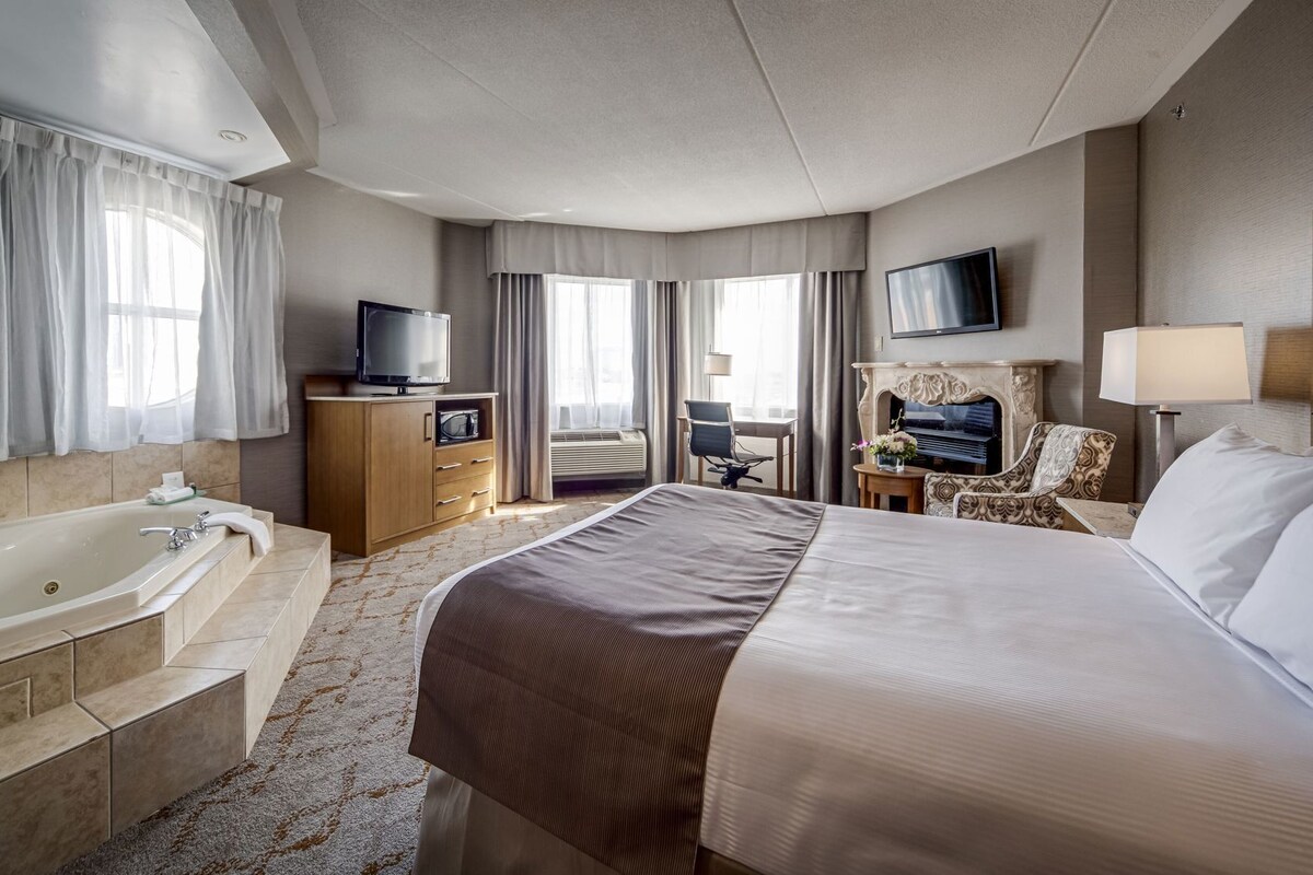 A hotel room with a large bed, a bathroom with a bathtub, a desk with a chair, a television, and a fireplace.