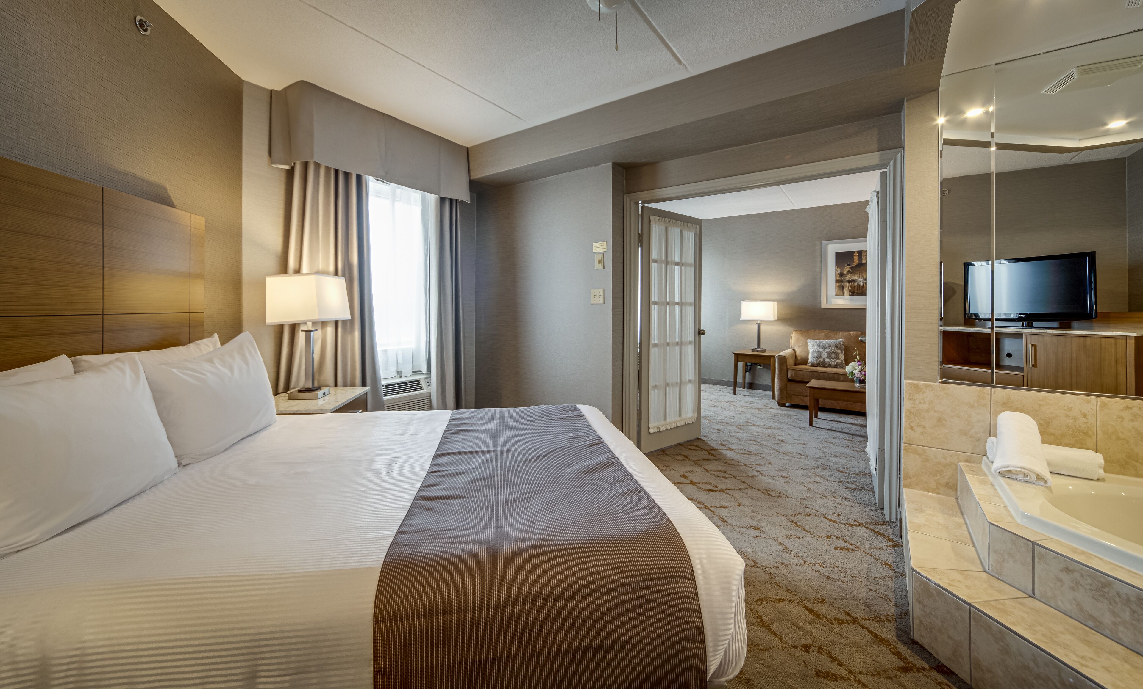 Quarto de hotel com uma cama grande, área de estar com sofá e TV, banheiro com banheira e corredor que leva a outro quarto.