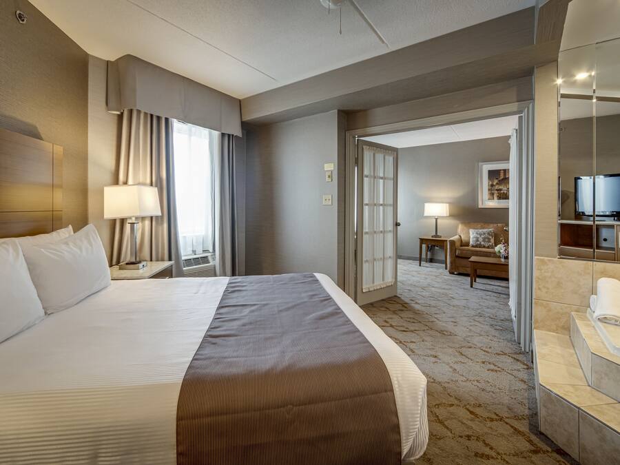 Habitación de hotel con una cama grande, una zona de estar con sofá y televisor, un baño con bañera y un pasillo que conduce a otra habitación.