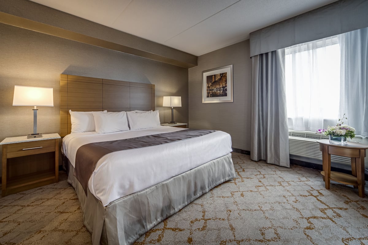 A hotel room with a large bed, two bedside tables with lamps, a framed picture on the wall, a window with curtains, and a small table with a vase of flowers.