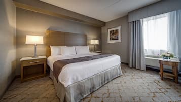 Habitación de hotel con una cama grande, dos mesitas de noche con lámparas, un cuadro en la pared, una ventana con cortinas y una pequeña mesa con un jarrón de flores.