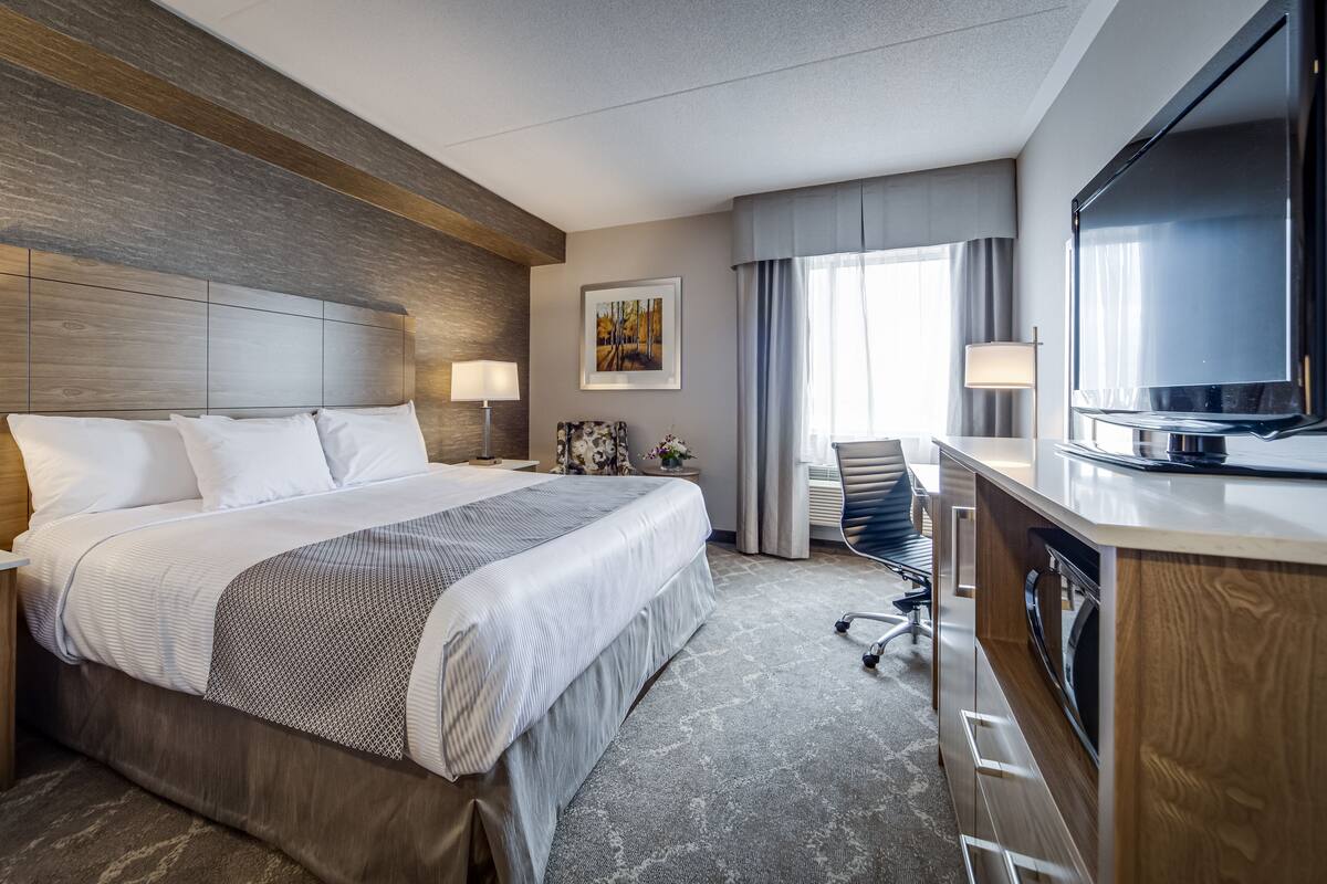 A hotel room with a large bed, a desk with a flat-screen TV, a chair, and a window with curtains.