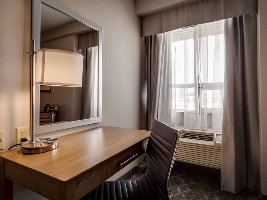 A hotel room with a wooden desk, a mirror, a lamp, a chair, a window with curtains, and an air conditioning unit.