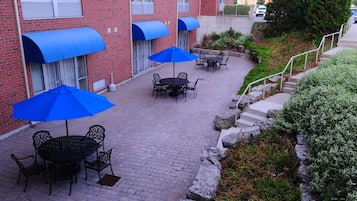 A brick building with outdoor seating areas featuring tables and chairs under blue umbrellas, a stone pathway, and a small garden area with rocks and shrubs.
