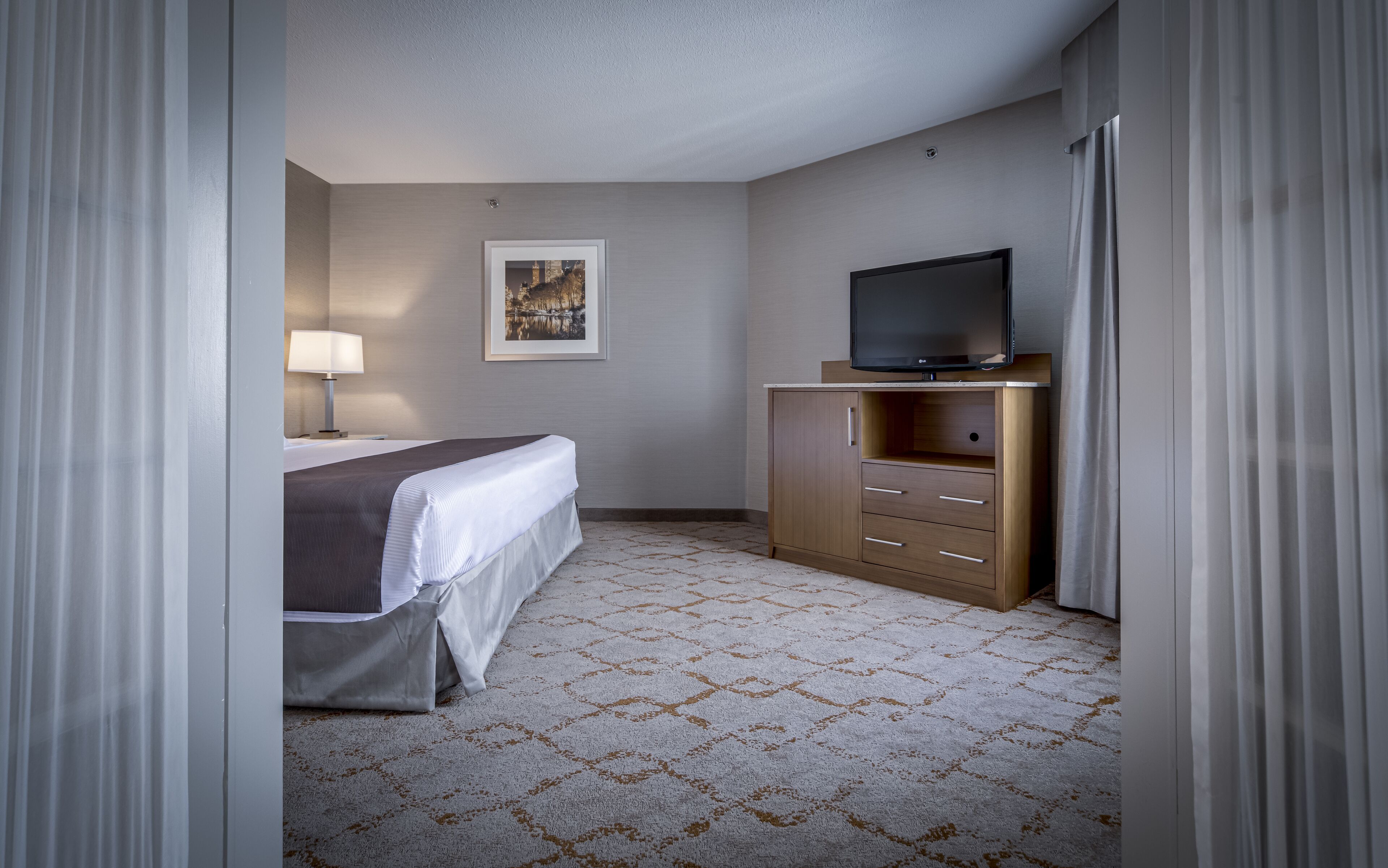 Une chambre d’hôtel avec un lit, un téléviseur posé sur une commode en bois, une lampe et un tableau encadré au mur.
