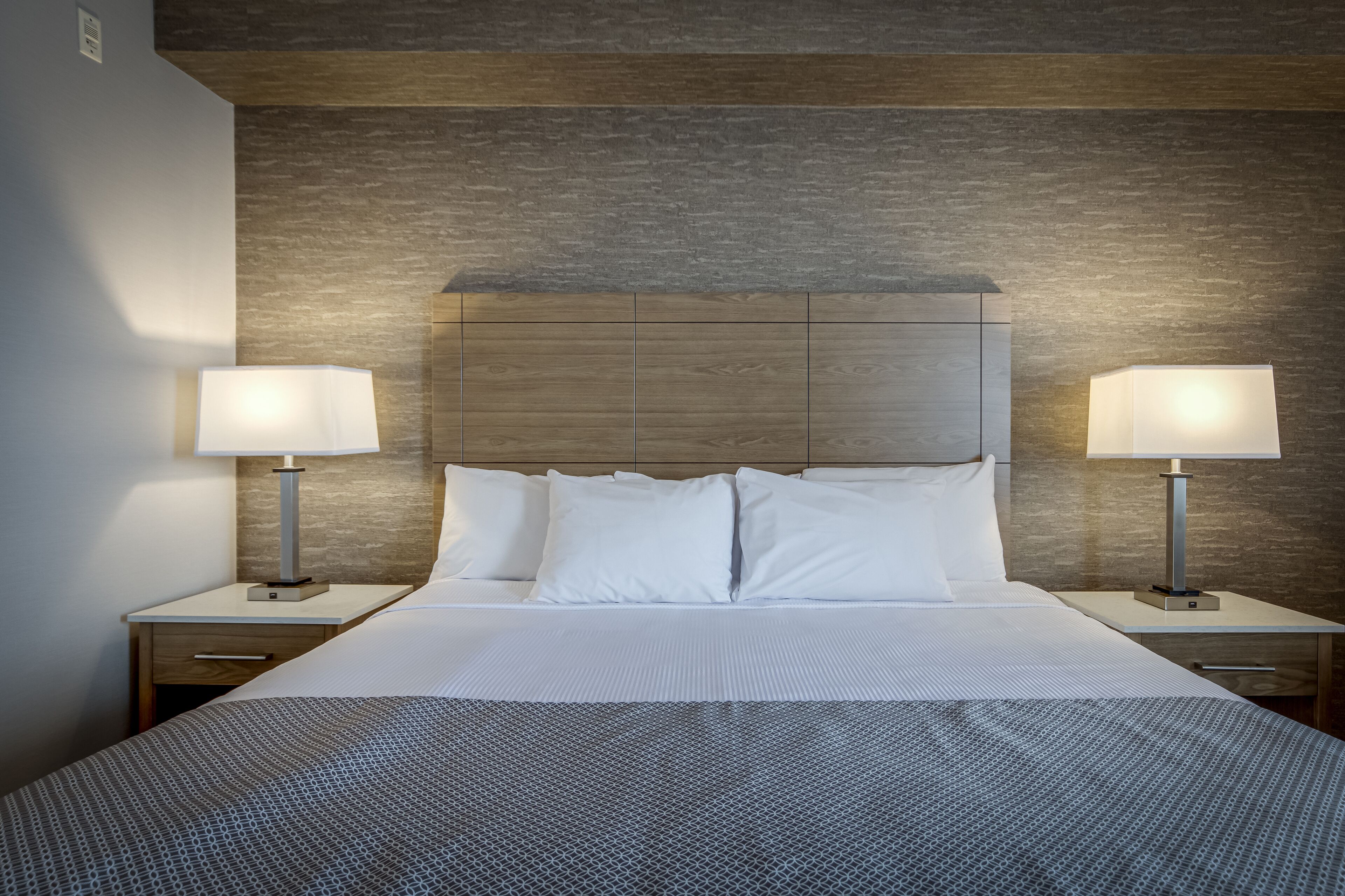 A neatly made bed with white linens and a patterned blanket, flanked by two bedside tables with lamps, set against a textured wall.
