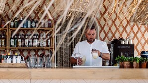 Un espace bar avec un comptoir, des étagères remplies de bouteilles et une machine Nespresso. Trois tabourets en osier sont placés devant le comptoir.